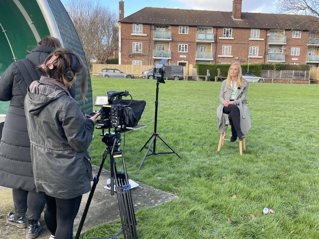Behind The Camera And Camera Operator Showing The Presenter Sat On A Stool In An External Filming Environment
